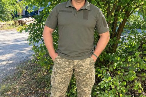 Чоловік в оливковому поло та камуфляжі стоїть біля дерев на стежці