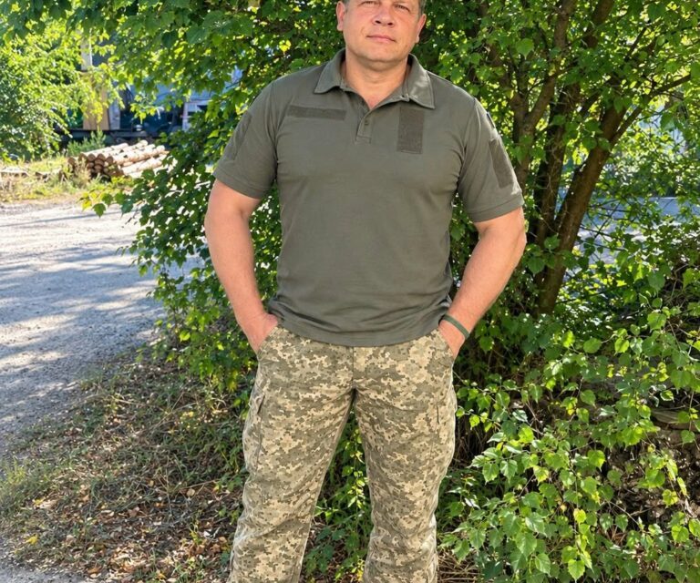 Чоловік в оливковому поло та камуфляжі стоїть біля дерев на стежці